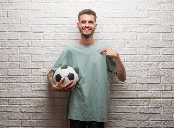 Молодий дорослий чоловік тримає футбольний м'яч, що стоїть над білою цегляною стіною з несподіваним обличчям, спрямованим пальцем до себе
 - Фото, зображення