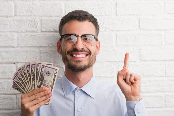 Joven hombre de negocios hipster sosteniendo dólares sorprendido con una idea o pregunta señalando el dedo con la cara feliz, número uno
 - Foto, imagen