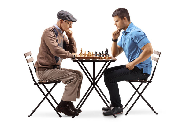 Senior and a young man seated at a table playing chess isolated on white background - Фото, зображення