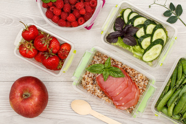 Apple and plastic meal prep containers with fresh strawberries, raspberries, boiled buckwheat porridge and slices of meat, cucumbers and salad, green peas, wooden spoon on grey background. Top view - Photo, Image