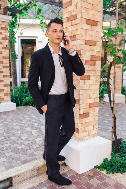 Young stylish handsome man posing in modern city park, wearing suit and sunglasses. - Фото, изображение