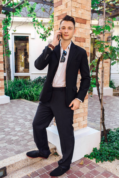 Молодой стильный красивый мужчина позирует в современном городском парке, носит костюм и солнцезащитные очки
. - Фото, изображение