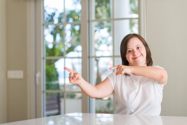 Down syndrome woman at home smiling and looking at the camera pointing with two hands and fingers to the side. - Photo, Image