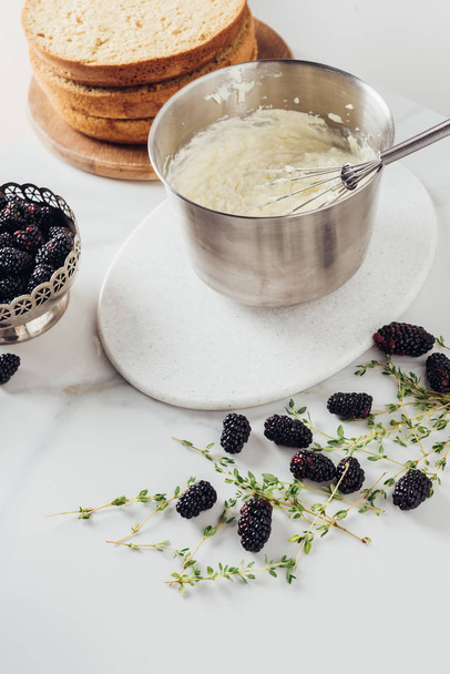 cropped shot of metal bucket with whisk and cream for cake and blackberries on white table - 写真・画像