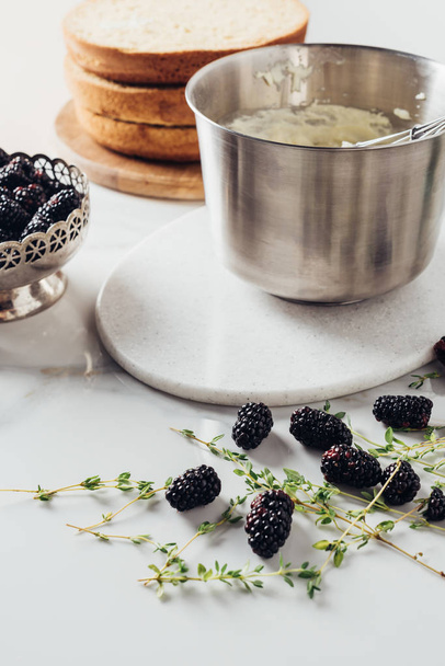 cropped shot of metal bowl with whisk and cream for cake and blackberries on white table - Фото, изображение