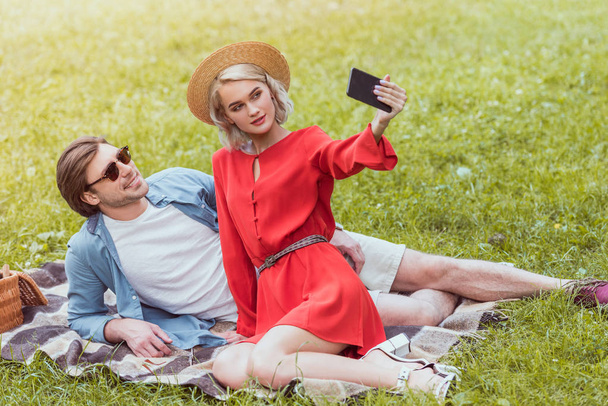 young couple sitting on blanket in park and taking selfie with smartphone - Foto, Imagen