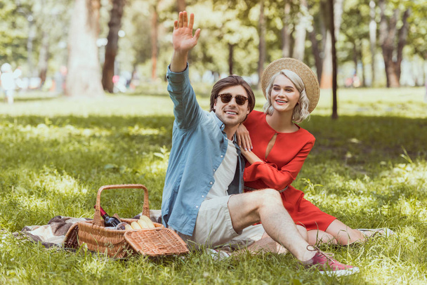 boyfriend sitting on blanket in park and waving hand - Photo, Image