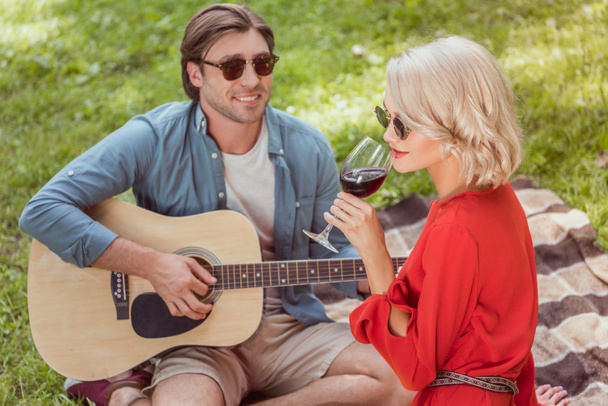 handsome boyfriend playing acoustic guitar, girlfriend drinking wine at picnic - Foto, Imagen