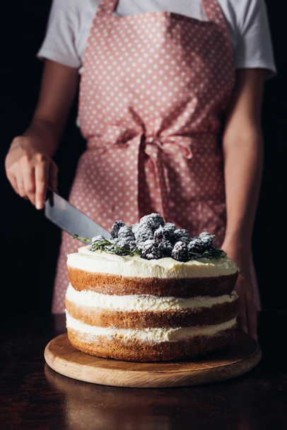 cropped shot of woman in pink apron cutting delicious blackberry cake on black - Zdjęcie, obraz