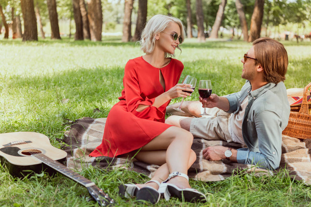 feliz pareja acostada en la manta en el parque y tintineo con copas de vino tinto
 - Foto, Imagen