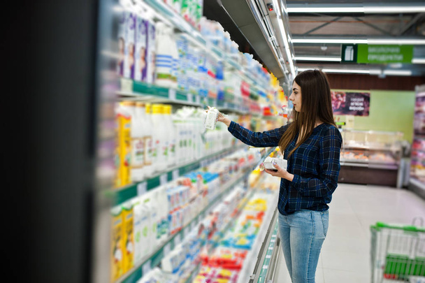 Γυναίκα ψάχνει στα ράφια του σούπερ μάρκετ για ψώνια. Πορτρέτο του ένα νεαρό κορίτσι σε ένα κατάστημα αγοράς εκμετάλλευση παραγωγής γάλακτος. - Φωτογραφία, εικόνα