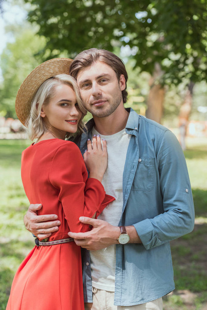 smiling girlfriend and boyfriend hugging in park and looking at camera - Фото, изображение