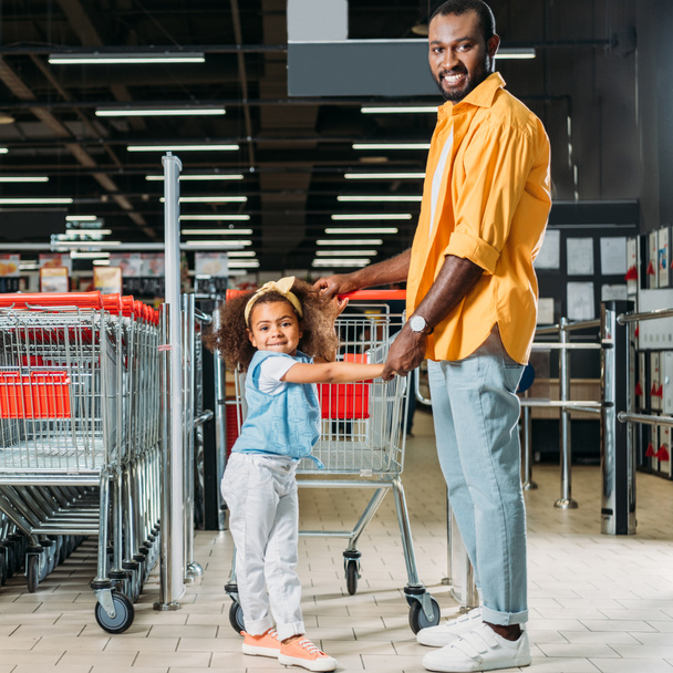 sonriente afroamericano hombre de pie con hija cerca de carrito de compras en el supermercado
  - Foto, Imagen