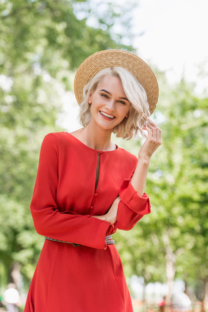smiling attractive girl in red dress and straw hat looking at camera in park - Fotoğraf, Görsel