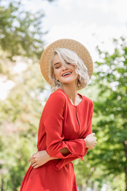 smiling attractive girl in red dress and straw hat in park - Foto, afbeelding