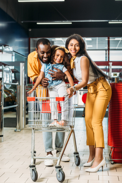 happy young african american couple with little daughter standing in shopping trolley in hypermarket  - Foto, afbeelding