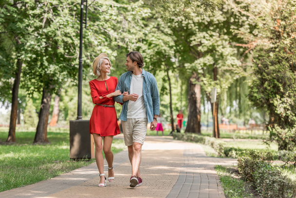 smiling girlfriend and boyfriend walking together in park and looking at each other - Фото, изображение