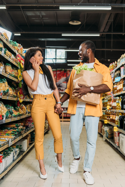 riant homme afro-américain avec sac en papier tenant la main de sa petite amie pendant qu'elle parle sur smartphone dans un supermarché
 - Photo, image