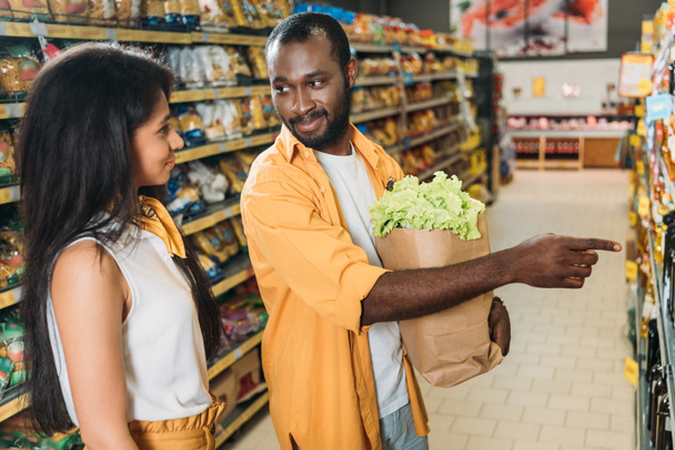 hombre afroamericano con bolsa de papel con comida apuntando a su novia en el supermercado
  - Foto, imagen