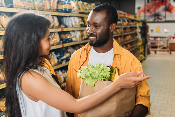 young african american woman pointing to boyfriend with paper bag with food in supermarket  - Foto, imagen