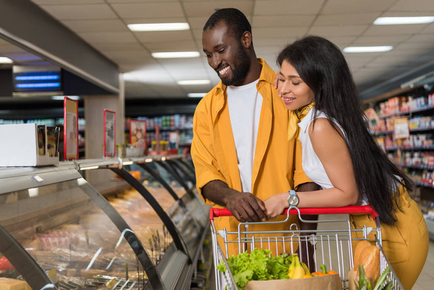  Lächelndes afrikanisch-amerikanisches Paar mit Einkaufswagen bei der Lebensmittelauswahl im Supermarkt  - Foto, Bild