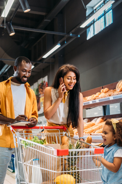 lächelnde Afroamerikanerin spricht auf Smartphone, während ihr Mann und ihre Tochter mit Einkaufswagen in der Nähe im Supermarkt stehen  - Foto, Bild