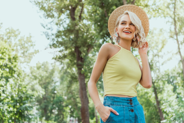 low angle view of stylish attractive blonde girl in straw hat looking away in park - Foto, Imagem