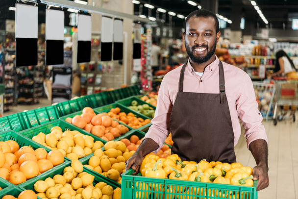 happy african american male shop assistant in apron holding box with bell peppers in grocery store - Fotó, kép