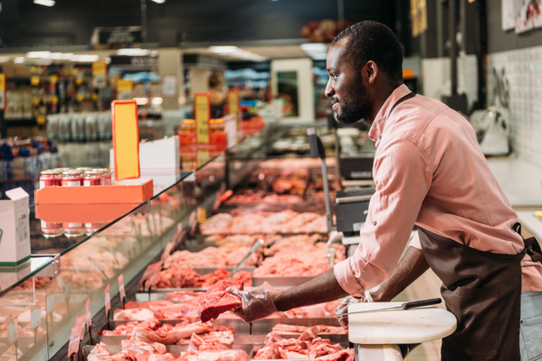 Afro-Amerikan erkek kasap süpermarkette biftek çiğ et veren önlük yan görünüm  - Fotoğraf, Görsel