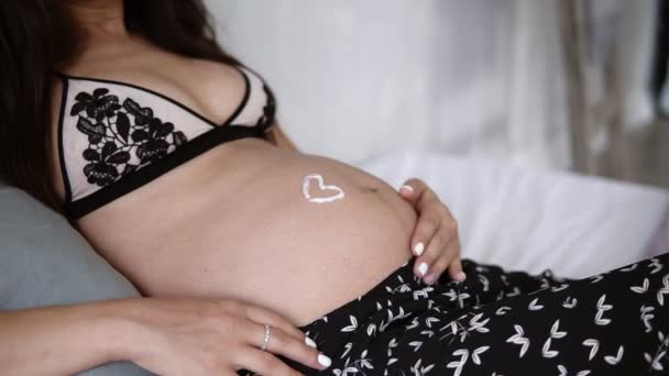 Close up of a lovely pregnant girl in beautiful bra lying on a bed with love symbol on the expectant mother stomach. Caressing her her belly with a cream - Filmati, video