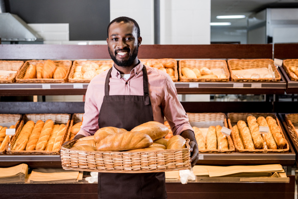selective focus of african american male shop assistant in apron with loaves of bread in supermarket  - Foto, afbeelding