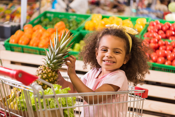 bambino afroamericano sorridente seduto nel carrello della spesa con ananas nel negozio di alimentari
  - Foto, immagini