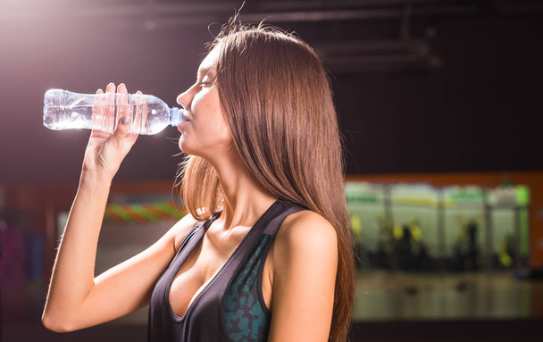 Фітнес-леді п'є воду з пляшки. М'язиста молода жінка в спортзалі робить перерву від тренувань
. - Фото, зображення