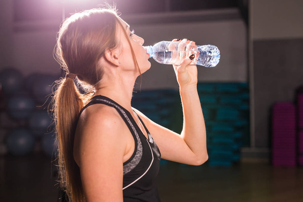 Фітнес-леді п'є воду з пляшки. М'язиста молода жінка в спортзалі робить перерву від тренувань
 - Фото, зображення