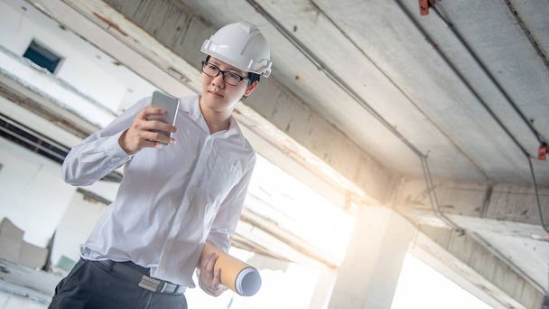 Asian male engineer or architect holding blueprints or architectural drawing while using smartphone for calling at construction site. Engineering, Architecture and building construction concepts - Foto, Imagem