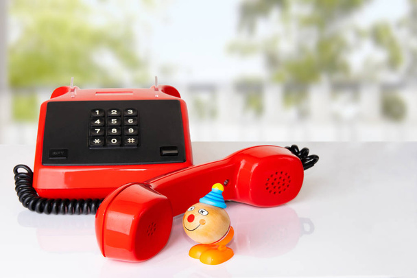 Red vintage telephone receiver with black cable on white table in front of natural blurred green background. Funny toy clown is taking a call. Suitable for contact exchange and call center. Template for your product display montage.  - Foto, immagini