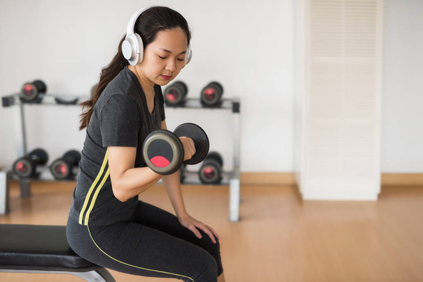 Fitness Asian woman work out with dumbbell and listen music by earphone at gym. Healthy girl has intensive training and hold sport equipment. Arm and hand building. Sport lifestyle concept. - Photo, Image