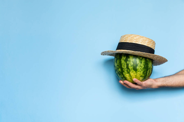 Man's hand holding a watermelon covered with straw hat. Light blue background. - Fotó, kép