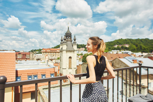attractive happy smiling woman walking in the center of old city of Lviv. woman admires the view of the cityscape from the rooftop with wonderful cloudy blue sky on background - 写真・画像