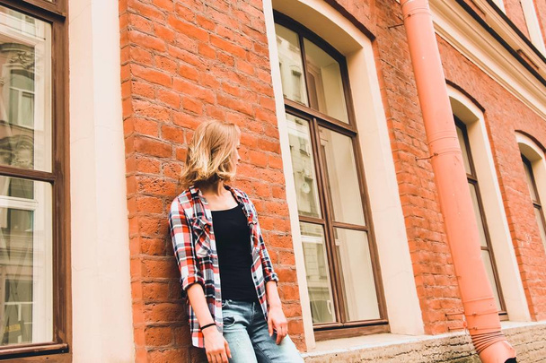 A beautiful and young girl stands near a brick house with large wooden windows. Her hair is loose and her head is turned to the left so that her face can not be seen. - Foto, Imagen