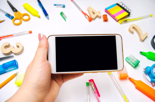 The student is holding a mobile phone in the white background. concept of education. back to school. creative mess on the desktop. desk. stationery. smartphone. place for text. flat lay. - Foto, afbeelding