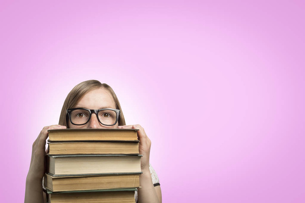 Юная студентка в очках выглядывает из-под груды книг на розовом фоне
. - Фото, изображение