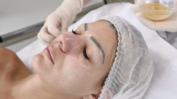 Conceito de beleza. Jovem cliente feminina recebe procedimento facial de beleza. renovação facial. Esteticista usa botão de algodão para cobrir a pele do rosto com máscara de beleza
 - Filmagem, Vídeo