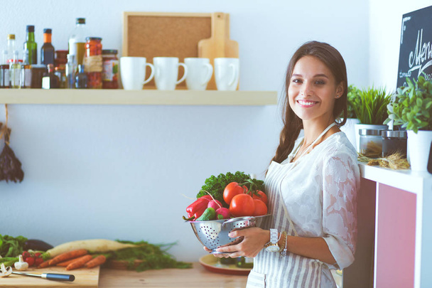 Smiling young woman holding vegetables standing in kitchen. Smiling young woman - Fotó, kép