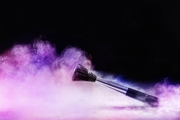 Makeup brush in colorful dust fog from powder explosion isolated on black background. Cosmetic and beauty art concept. Close up, selective focus - Photo, Image