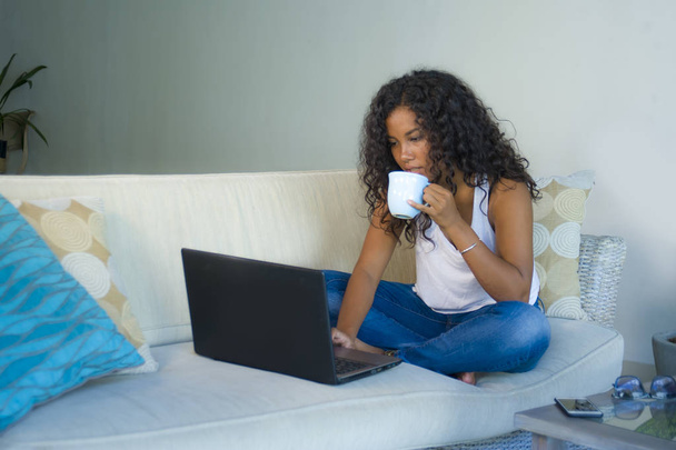 lifestyle portrait of young attractive and relaxed latin American student woman sitting at home sofa couch networking with laptop computer drinking coffee or tea wearing jeans and singlet - Photo, image