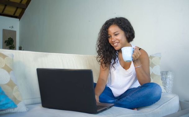 lifestyle portrait of young attractive and relaxed latin American student woman sitting at home sofa couch networking with laptop computer drinking coffee or tea wearing jeans and singlet - Zdjęcie, obraz