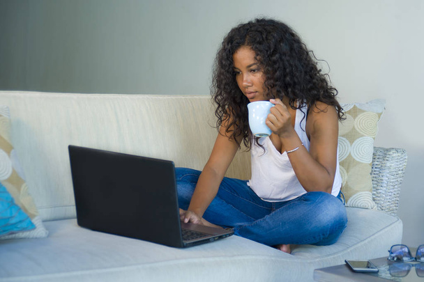 lifestyle portrait of young attractive and relaxed afro American student woman sitting at home sofa couch networking with laptop computer drinking coffee or tea wearing jeans and singlet - Foto, Bild