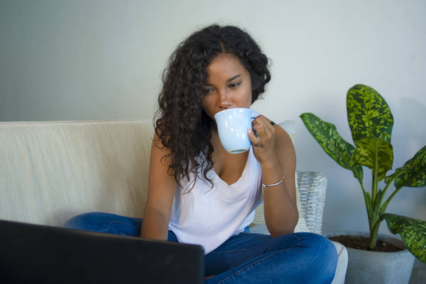lifestyle portrait of young attractive and relaxed black afro American student woman sitting at home sofa couch networking with laptop computer drinking coffee or tea wearing jeans and singlet - Zdjęcie, obraz