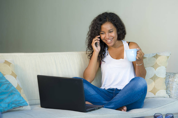 lifestyle isolated portrait of young happy and beautiful black african American woman talking on mobile phone while working on laptop computer at home living room sofa couch  - Photo, Image